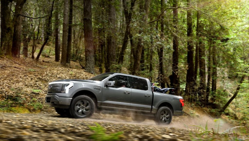 Profile view of a 2022 Ford F-150 Lightning being driven on a road in a heavily wooded area. | Ford dealer in Jacksonville, AR.